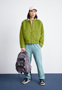 Green fuzzy jacket with button closure, light beige top, mint green sweatpants, gray backpack with straps, and black/white sneakers.