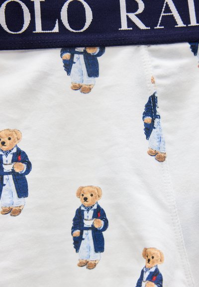 Λευκό ύφασμα με επαναλαμβανόμενα αρκουδάκια που φοράνε μπλε ρόμπες και κρατούν φλιτζάνια τσαγιού, και μπλε σκούρο ζωνάρι μέσης με λευκό κείμενο.