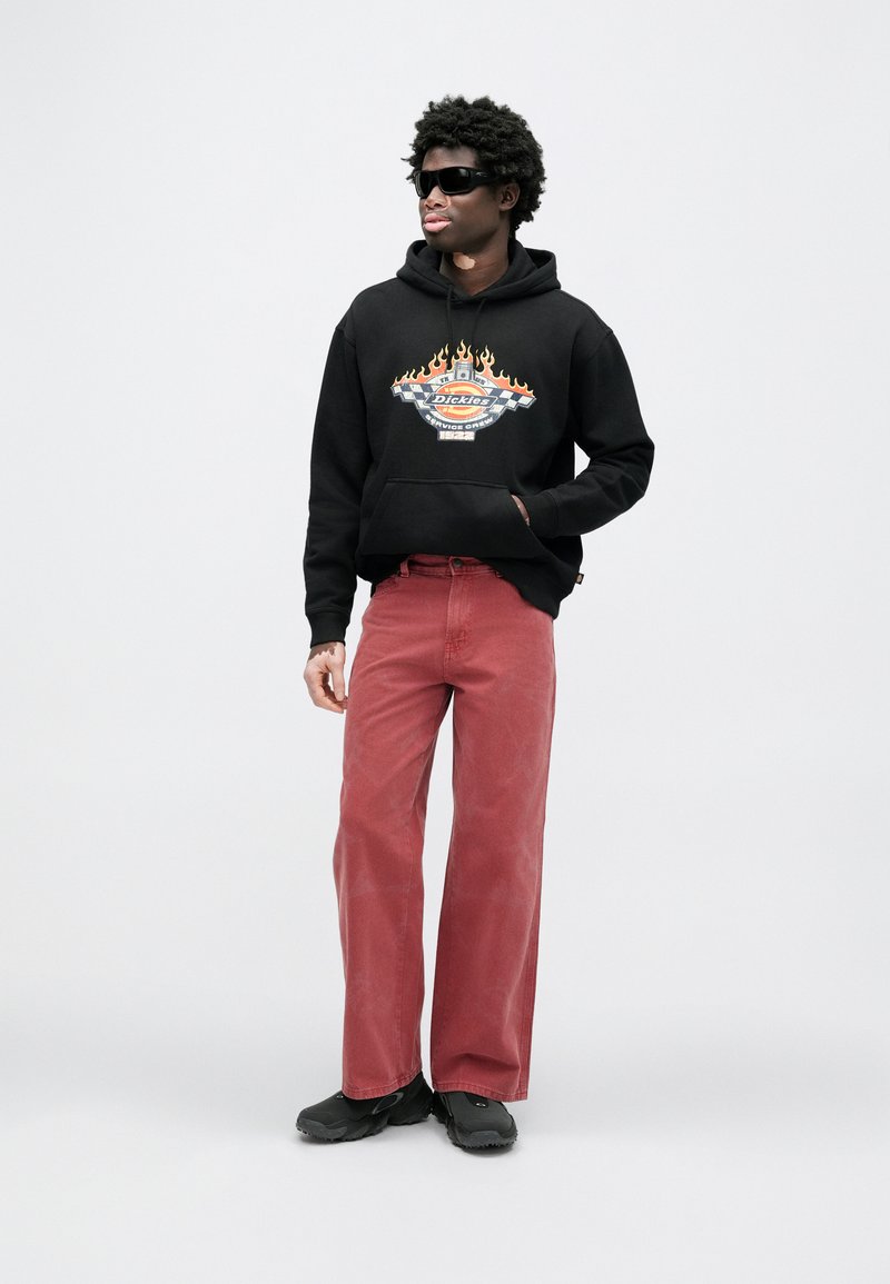 Black hoodie with flame graphic and logo, red wide-leg pants, and black footwear. Model wears dark sunglasses against a light background.