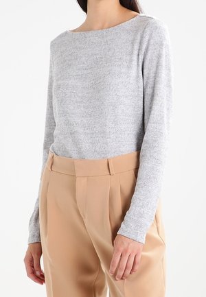 Kobieta ubranа w jasnoszary top z długim rękawem, wpuszczony w spodnie beżowe o wysokim stanie z szlufkami na pasek, stojąca na białym tle.