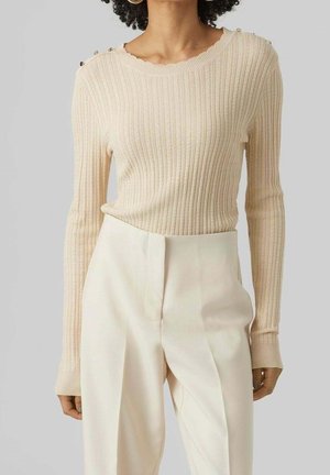 Vrouw in een beige geribde trui met lange mouwen en schouderknopen, gecombineerd met hooggerande crèmekleurige pantalon, tegen een grijze achtergrond.