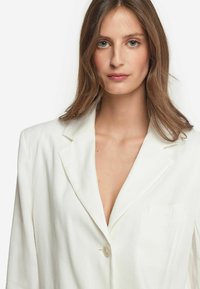 Vit blazer i lätt tyg. Har en snedskuren krage, enkel knappstängning och en bröstficka. Slät textur, skräddarsydd design.