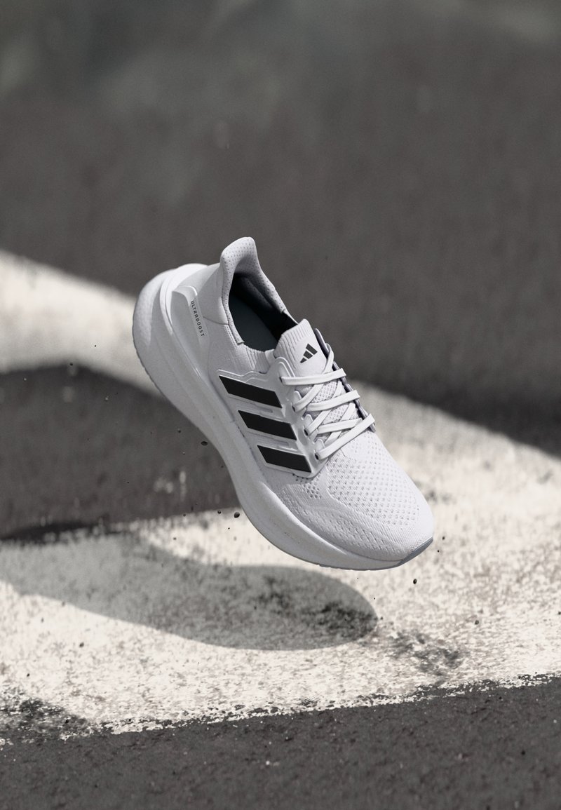 Valkoinen urheilukenkä, jossa on hengittävä verkkopäällinen, mustat raidat, pehmustettu kaulus ja pehmeä pohja. Kelluu maalatun viivan yläpuolella.