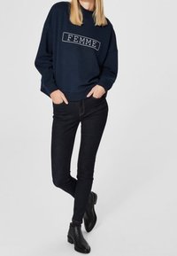 Felpa blu navy con la scritta "FEMME", abbinata a jeans skinny neri e stivali neri a caviglia, su uno sfondo semplice.