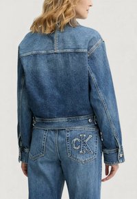 Kurtka dżinsowa w średnim niebieskim kolorze o klasycznym kroju, z mankietami zapinanymi na guziki, bocznymi kieszeniami oraz wyszywanym logo „CK” na tylnej kieszeni.