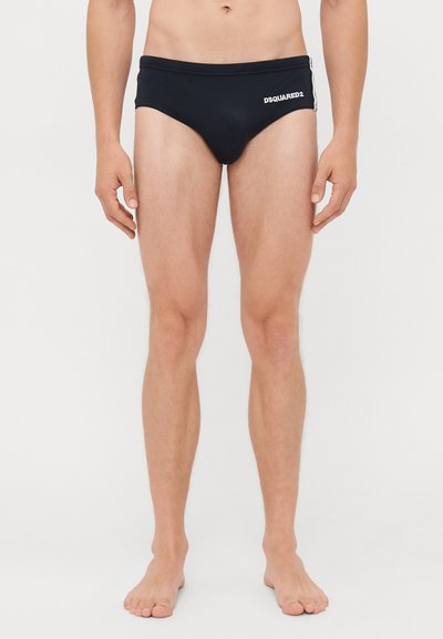 Uomo che indossa slip da bagno neri DSQUARED2, in piedi a piedi nudi contro uno sfondo bianco.