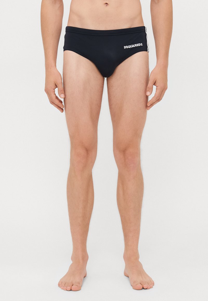 Mann in schwarzen DSQUARED2 Badebriefs, barfuß vor weißem Hintergrund stehend.