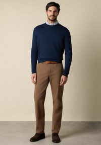 Marineblauer Rundhals-Pullover kombiniert mit braunen Chinos, ergänzt durch einen braunen Gürtel und dunklen Schuhen, in einem minimalistischen Umfeld mit beigem Hintergrund.