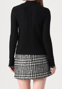 Femme portant un haut noir côtelé à manches longues ajusté et une mini-jupe à motif pied-de-poule noir et blanc avec une fermeture éclair arrière visible.