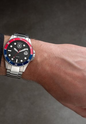 Montre-bracelet avec un bracelet en métal argenté, un cadran noir, une lunette rouge et bleue, et un affichage de la date indiquant "22" au poignet d'une personne.