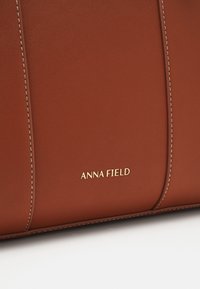 Section d'un sac en cuir marron avec des coutures verticales et un petit logo doré "Anna Field" sur une surface lisse.