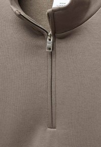 T-shirt zippé marron avec un col haut, texture côtelée et tirette en métal gravée du logo de la marque. Doublure intérieure douce visible.