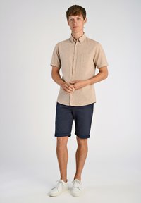 Camicetta a maniche corte di un leggero colore beige con taschino sul petto, abbinata a pantaloni corti blu navy arrotolati e sneakers bianche. Design semplice e casual.