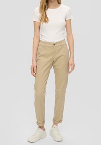 Femme portant un haut blanc à manches courtes, un pantalon beige taille haute retroussé, et des baskets blanches, se tenant devant un fond uni.