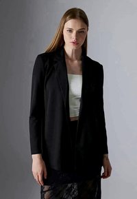 Νεαρή γυναίκα με μακριά μαλλιά φοράει μαύρο σακάκι πάνω από λευκό τοπ και μαύρη δαντελένια φούστα, στέκεται μπροστά σε απλό γκρι φόντο.