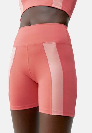 Korte sportieve shorts in een koraaltint, uitgevoerd in een gladde stof met een contrasterend doorzichtig paneel langs de zijkanten. Strak model met een hoge tailleband.