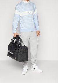 Světle modrý a bílý pruhovaný polo s dlouhým rukávem, šedé tepláky a bílé tenisky. Černá sportovní taška s logem "Champion", texturovaná tkanina.