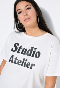 Biała koszulka z krótkim rękawem z napisem "Studio Atelier" w pogrubionym, czarnym druku. Miękka tkanina o luźnym kroju i okrągłym dekolcie.
