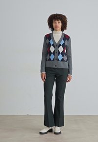 Grå cardigan med fargerikt argyle-mønster i blått, rødt og hvitt. Har V-hals, knapper foran og er kombinert med svarte utsvingte bukser.