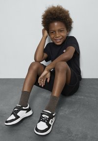 T-shirt noir et short portés avec des baskets noires et blanches à sangle Velcro, bout rond et semelle texturée. Des chaussettes brunes complètent le look.