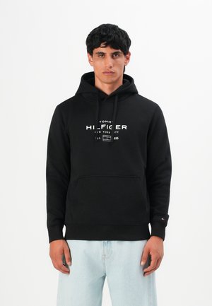 Uomo che indossa un hoodie nero di Tommy Hilfiger con logo bianco e jeans azzurri chiari, in piedi contro uno sfondo bianco semplice.
