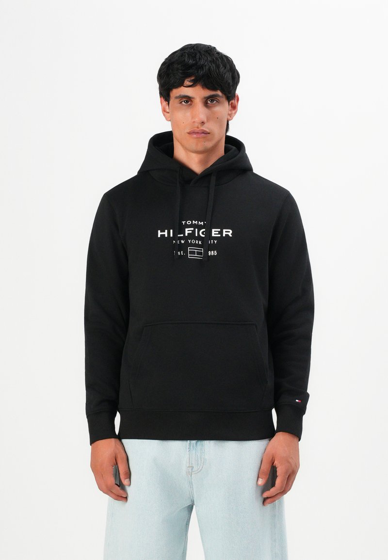 Hombre vestido con una sudadera negra de Tommy Hilfiger con texto en logo blanco y jeans de color azul claro, de pie contra un fondo blanco liso.
