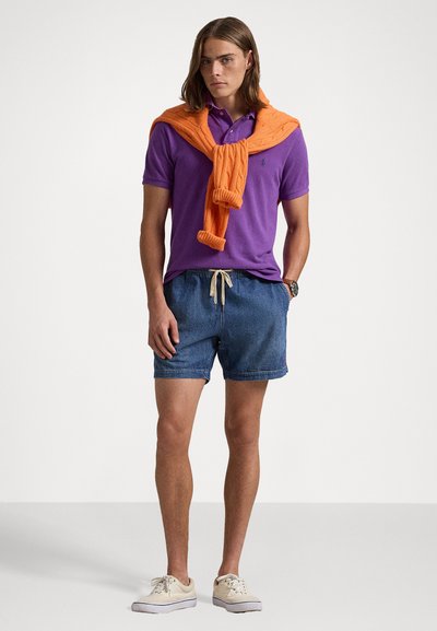 Lilla polo skjorte med korte ermer, en oransje strikket genser knyttet rundt halsen, denimshorts og lyse sneakers.