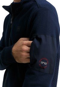 Granatowa kurtka zapinana na zamek z teksturowanymi ramionami, z naszywką z logo na rękawie i gładką tkaniną, nadającą elegancki wygląd.