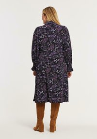 Frau in einem schwarzen Midikleid mit lila Blumenmustern und braunen Wildleder-Stiefeln in Knielänge mit Absatz, die mit dem Rücken zu einem schlichten Hintergrund steht.