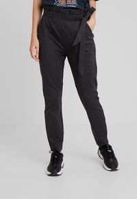 Tmavě šedé zúžené kalhoty s papírovým pasem, uvázané na uzel na boku, nošené s černobílými sportovními botami a zastrčeným tričkem s grafikou.