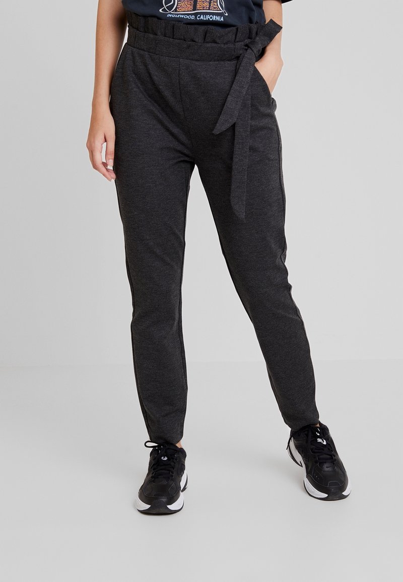 Tmavě šedé zúžené kalhoty s papírovým pasem, uvázané na uzel na boku, nošené s černobílými sportovními botami a zastrčeným tričkem s grafikou.