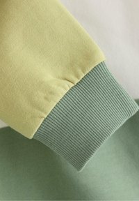 Ściągacz bluzy w jasnym żółtym kolorze z prążkowanym zielonym wykończeniem, prezentujący miękką teksturę i kontrastowe kolory w warstwowym designie.