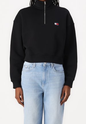 Person wearing a black half-zip sweatshirt with a small Tommy Jeans logo and light blue high-waisted denim jeans.