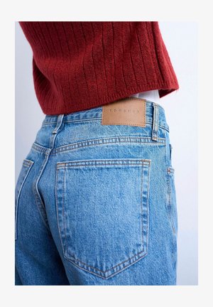 Persoon draagt lichtblauwe denim Topshop jeans en een rode geribbelde trui, gefotografeerd van de taille naar beneden, achteraanzicht.