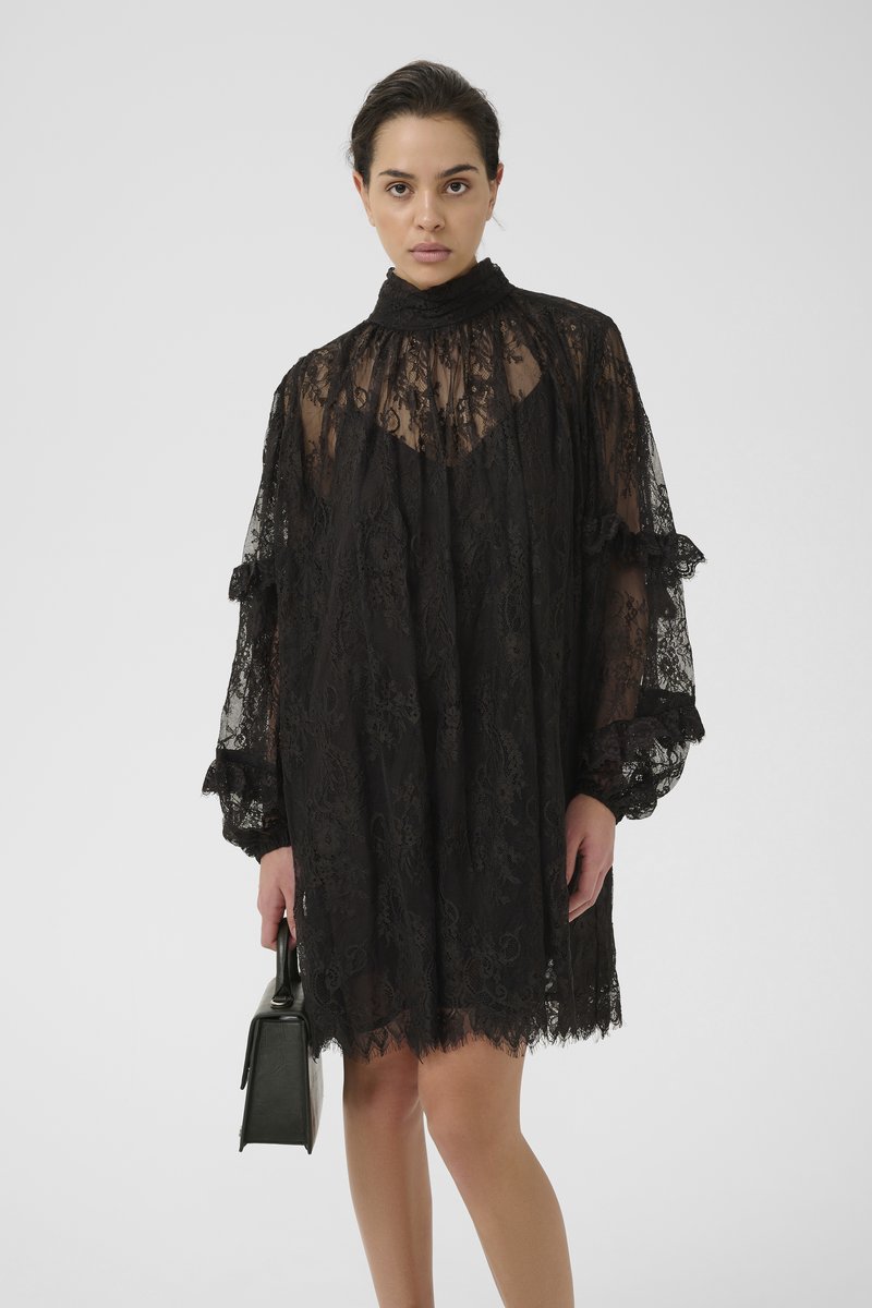 Black lace dress with a high neck, puffed sleeves, and ruffled accents. The sheer fabric features floral patterns and a loose fit. Holds a black handbag.