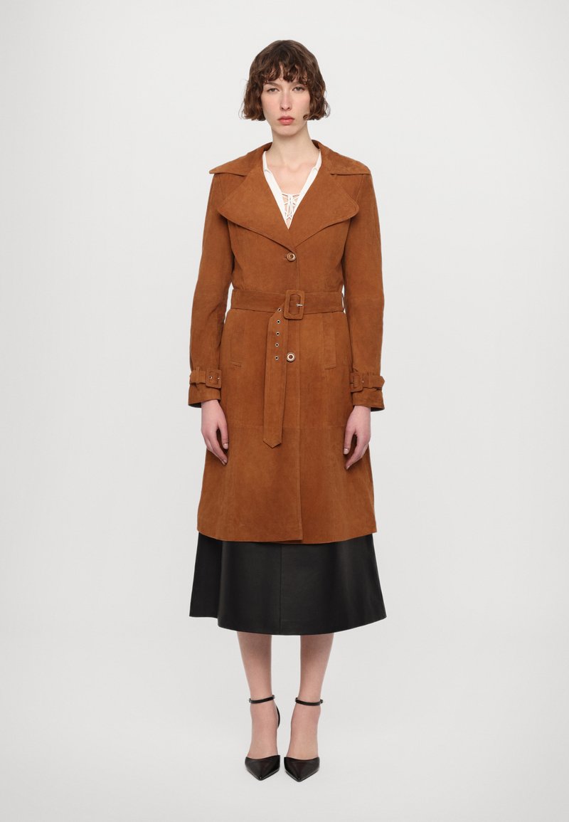 Trench coat in suede marrone con ampio colletto, vita con cintura e dettagli con bottoni. Indossato sopra una gonna nera, abbinato a scarpe nere con punta.