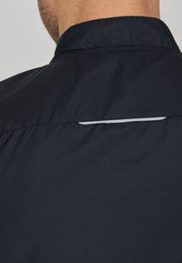 Hemd aus leichtem, navyblauen Stoff mit Texturmuster und reflektierenden Akzenten am hinteren Ausschnitt. Das Material wirkt glatt und atmungsaktiv.