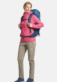 Veste magenta à fermeture éclair avec capuche, pantalon beige clair et un sac à dos bleu. Chaussures robustes et sangles de matériel visibles. Texture lisse et design sportif.