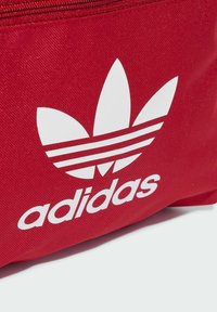 Bolsa de tela roja con un gran logotipo blanco de Adidas que presenta tres formas parecidas a hojas y dos franjas horizontales, con una superficie texturizada.