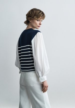 Woman with short wavy hair wearing a navy blue striped vest over a white long-sleeve blouse and white pants, looking over her shoulder.