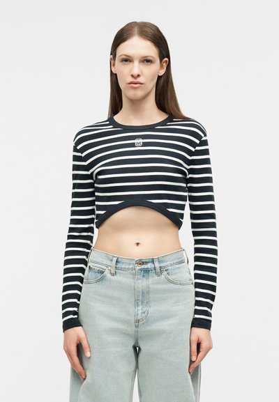 Νέα γυναίκα που φοράει μακρυμάνικο τοπ με μαύρες και λευκές ρίγες και ανοιχτόχρωμο τζιν ψηλόμεσο, στεκόμενη μπροστά σε απλό φόντο.