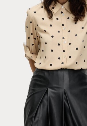 Chemise beige boutonnée à pois noirs portée rentrée dans une jupe plissée en cuir noir taille haute.