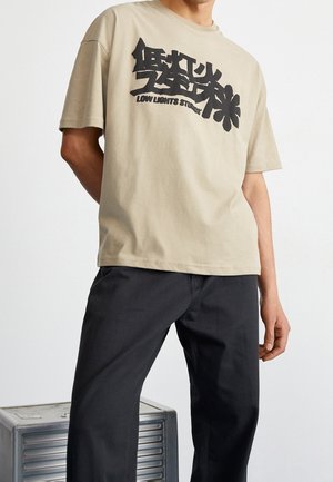 Mężczyzna ubrany w beżowy t-shirt z czarną abstrakcyjną grafiką i napisem "Low Lights Studio", w czarnych spodniach, stojący obok metalowej skrzynki.