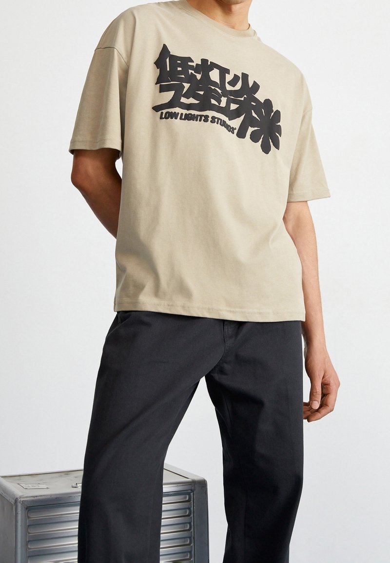 Homme portant un t-shirt beige avec un graphisme abstrait noir et le texte "Low Lights Studio", associé à un pantalon noir, debout près d'une boîte métallique.