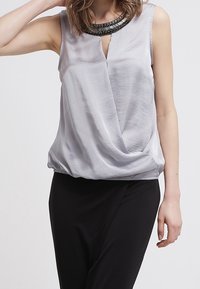 Blouse argentée sans manches avec un design drapé et un col en V. Dotée d'un col perlé et d'une texture de tissu douce et lisse. Portée avec un pantalon noir.