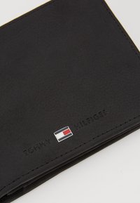Svart lær lommebok med en teksturert overflate. Har preget "TOMMY HILFIGER" tekst og en liten logo i rødt, hvitt og blått.