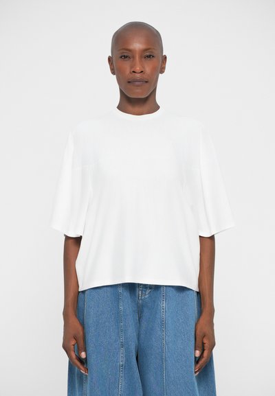 Eine glatzköpfige Person mit dunkler Hautfarbe, die ein locker sitzendes weißes Kurzarmhemd und weite blaue Jeans trägt, steht vor einem schlichten Hintergrund.