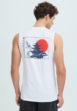 Homme portant un débardeur blanc avec un motif de pagode, deux grues et un soleil rouge dans le dos, debout devant un fond uni.