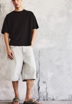 Homme portant un t-shirt noir ample, un short en jean gris clair au genou et des sandales grises à boucle, debout contre un mur gris texturé.
