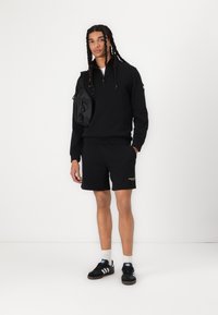 Zwarte fleece trui met een ritskraag, gecombineerd met zwarte shorts. Heeft een klein geel logo, zwarte schoudertas en zwarte sportschoenen.